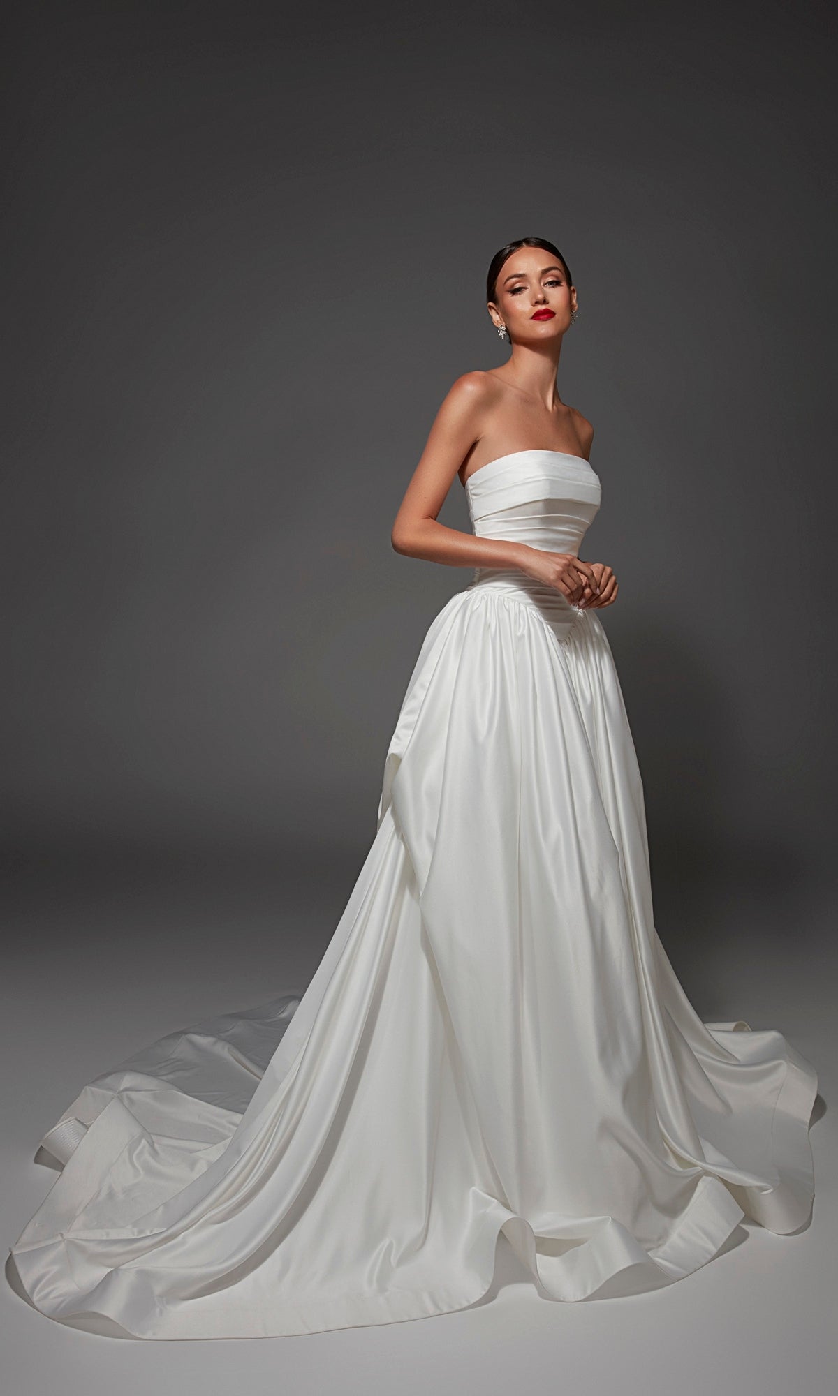 A woman wearing the Alyce 7187 long wedding dress—a strapless satin gown with a full skirt and long train—stands against a plain grey background.